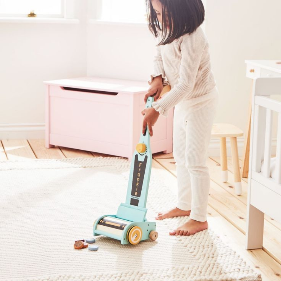 A little girl happily plays with a toy vacuum cleaner, smiling as she pretends to clean the floor.