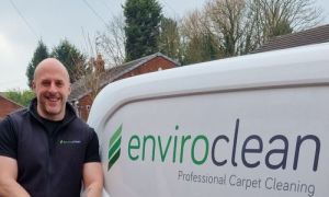 A smiling person stands by a white van labeled "enviroclean Professional Carpet Cleaning" with trees and a brick house in the background.