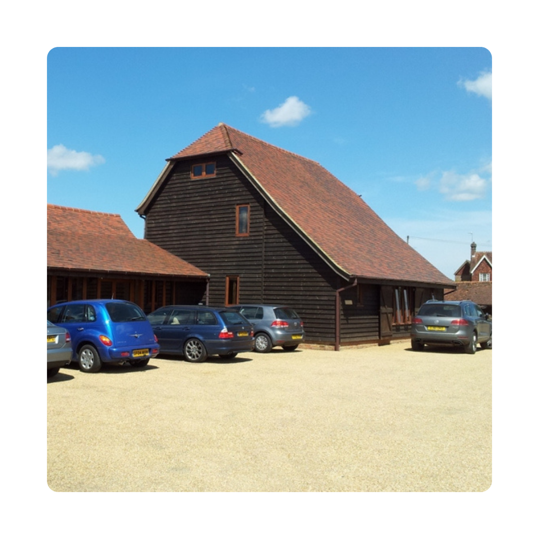 A rustic wooden barn with a red-tiled roof is surrounded by parked cars on a sunny day. Blue sky with a few clouds enhances the tranquil scene.