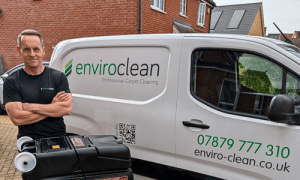 Man in a black shirt stands next to a white van labeled "enviroclean Professional Carpet Cleaning" with contact details, appearing professional and approachable.