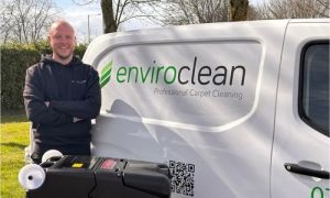 A smiling man in a hoodie stands beside a van with "enviroclean" branding. A carpet cleaning machine is in the foreground. The scene is bright and cheerful.