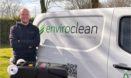A smiling man in a hoodie stands beside a van with "enviroclean" branding. A carpet cleaning machine is in the foreground. The scene is bright and cheerful.