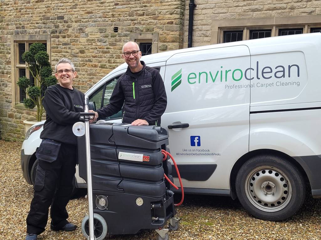 Two smiling people stand in front of an "Enviroclean" van with carpet cleaning equipment. They appear cheerful, conveying professionalism and teamwork.