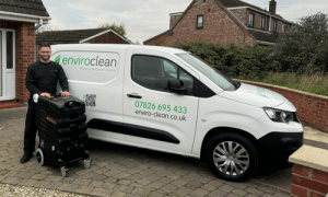 Man stands beside a white Enviroclean van holding cleaning equipment. The van displays contact info. Suburban house and overcast sky in background.