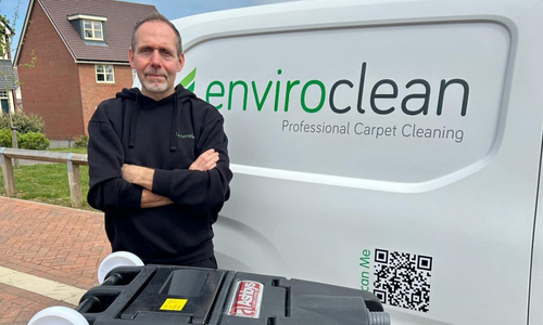 A person stands confidently in front of an Enviroclean van, showcasing carpet cleaning equipment. The scene is set outdoors beside a brick house.
