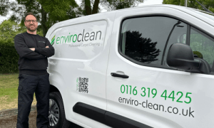 Man in black uniform leans against a white van labeled "enviroclean Professional Carpet Cleaning," set outdoors with green trees in the background.