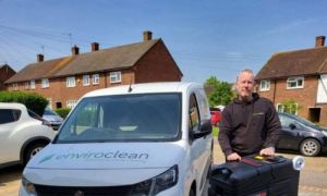 Man in a black hoodie stands next to a portable carpet cleaner and a white van with "Enviroclean" logo. Suburban houses and cars are in the background.