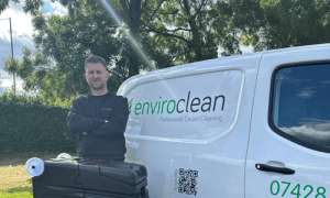 Man standing with arms crossed next to a white van labeled "enviroclean Professional Carpet Cleaning" in green and black text. Sunny outdoor setting.