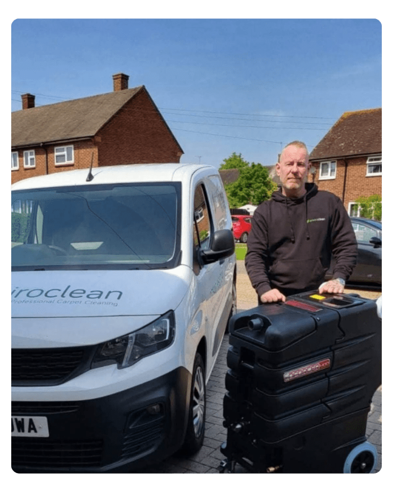 Man in a black hoodie stands next to a portable carpet cleaner and a white van with "Enviroclean" logo. Suburban houses and cars are in the background.