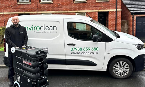 A smiling man stands beside a white "enviroclean" van with contact info, holding carpet cleaning equipment. The setting is a suburban street with brick houses.