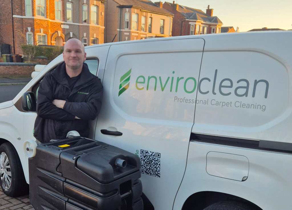 A person in a dark jacket leans against a white van with "enviro clean Professional Carpet Cleaning" written on the side. Residential street in the background.