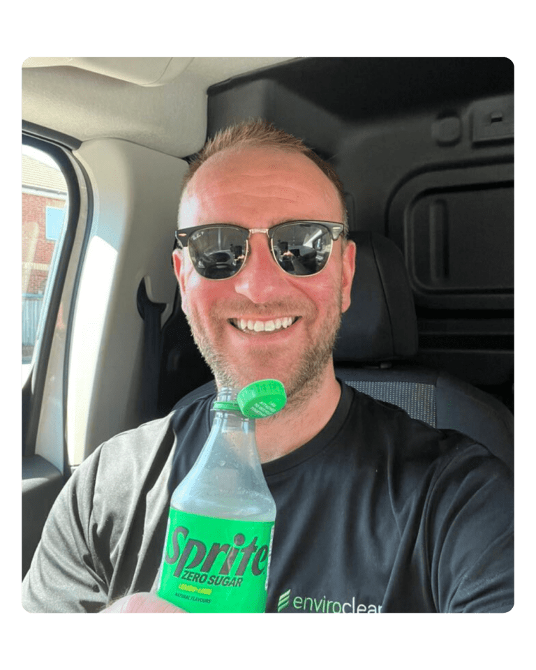 Man smiling in a vehicle, wearing sunglasses and a black "enviroclean" shirt, holding a bottle of Sprite Zero Sugar. Bright and cheerful mood.