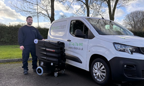 A smiling man stands beside a white Enviro clean van holding cleaning equipment. The scene is outdoors with trees and a bright sky.