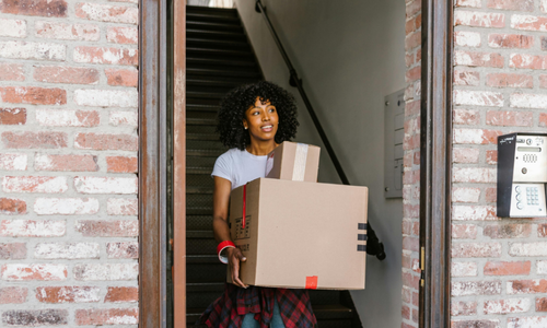 Smiling woman in a carpeted stair well doorway, walking out with stacked cardboard boxes. likely moving home.