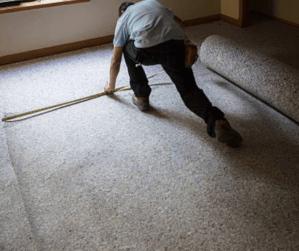 Technician putting down brand new grey carpet in a beige room with a window.