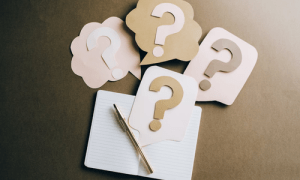 A desktop with a notebook open, but blank, with a pen and questions marks laid on top.