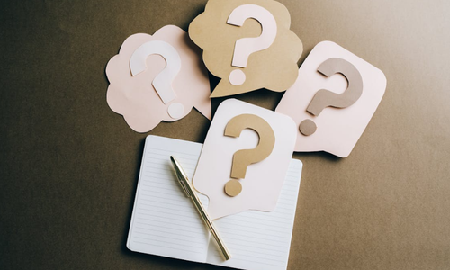 A desktop with a notebook open, but blank, with a pen and questions marks laid on top.