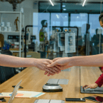 A board room full of people looking at paperwork, and laptops, with two people smiling over the desk at each other whilst shaking hands in agreement.