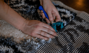 a pair of hands with a rug making loop tool, weaving fibres into a trellis, making a cream, blue and beige wool loop rug.